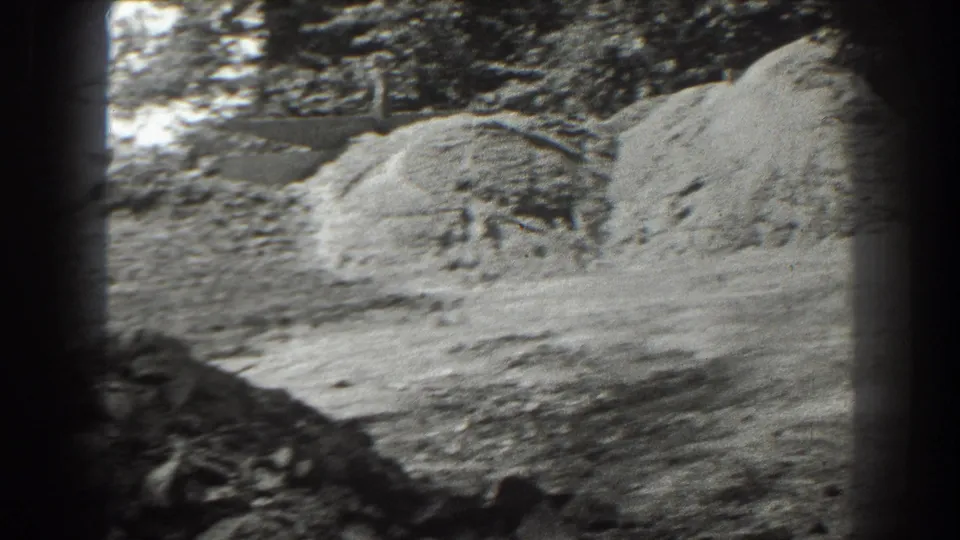 A Cumberland work yard in 1938 appears with mounded sand or ore, a dirt path, trees, and soft black-and-white 8mm texture.