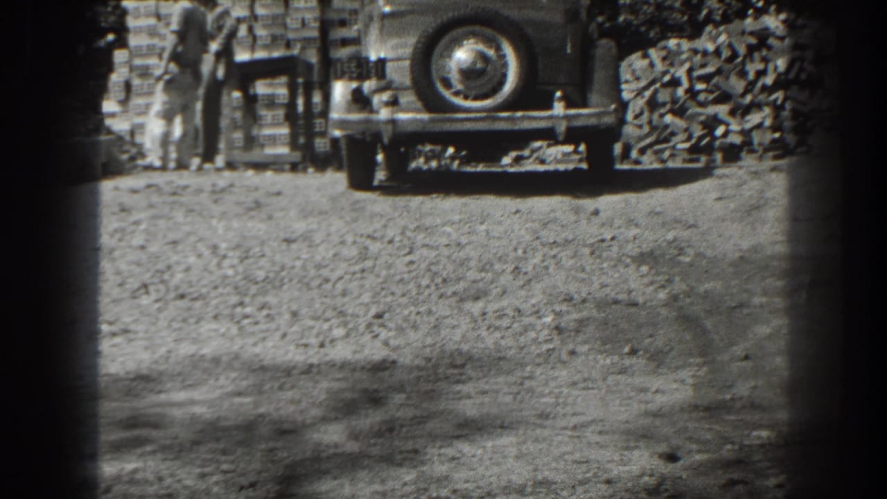 Two men stand near stacked bricks and the rear of a parked car on a dirt surface.