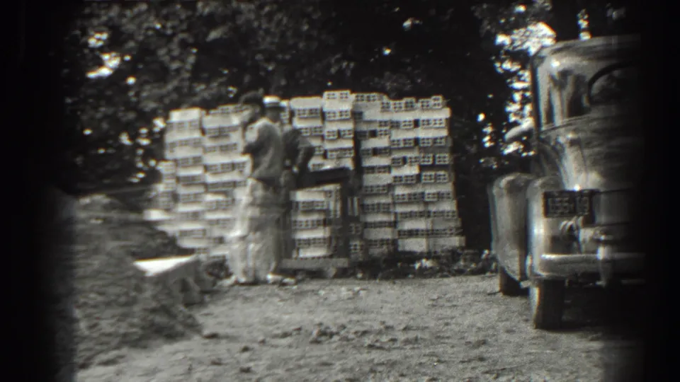 Two men in work clothes stand in conversation near stacked bricks and a parked car in the 1938 Cumberland home-movie scene.