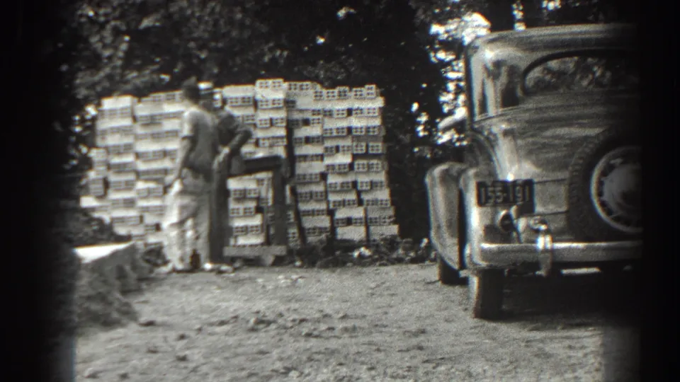 A man remains beside the brick stack as the rear of the 1930s car, including a visible plate, frames the Cumberland street-side scene.