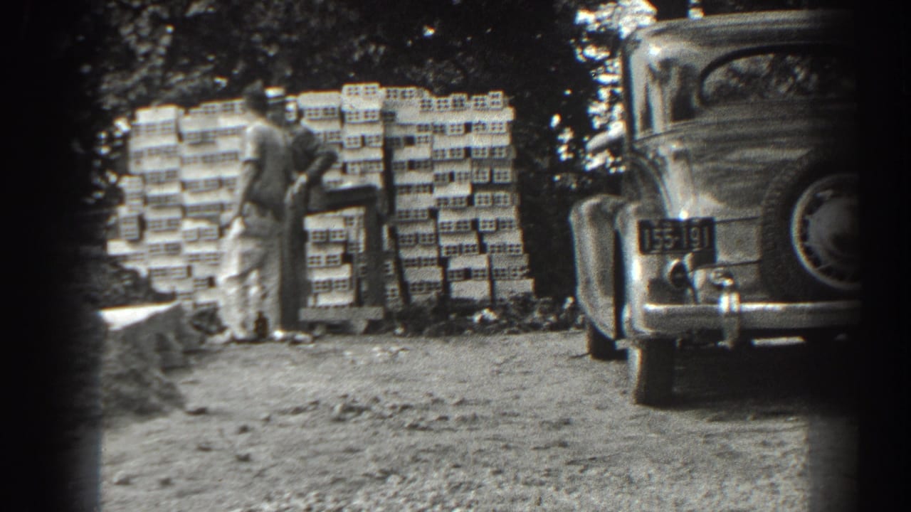 Two men stand near stacked supplies beside a parked vintage car on a dirt work site.
