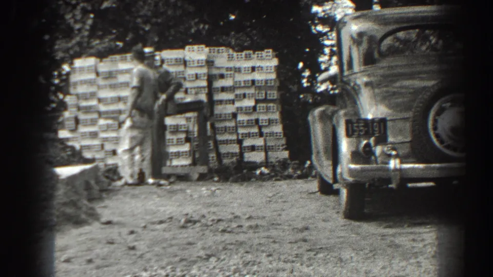 In 1938 Cumberland, Maryland footage, two men stand near a large stack of supplies beside a parked vintage automobile on a dirt lot.