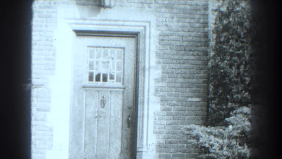 1938 Cumberland, Maryland 8mm frame showing a closed wooden door, divided glass window, pale trim, brick wall, and shrub at right.
