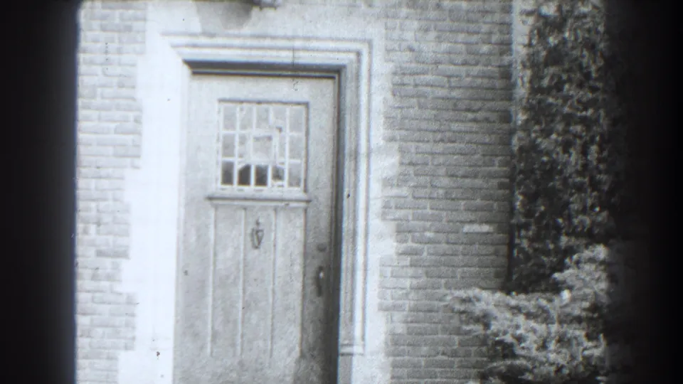 Softly focused 1938 Cumberland frame of a closed wooden door with divided glass, pale surround, brick wall, and shrub to the right.