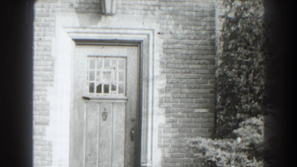 1938 Cumberland, Maryland home-movie frame showing a brick house entrance with a paneled wooden door, small grid window, ivy, and garden shrubs.
