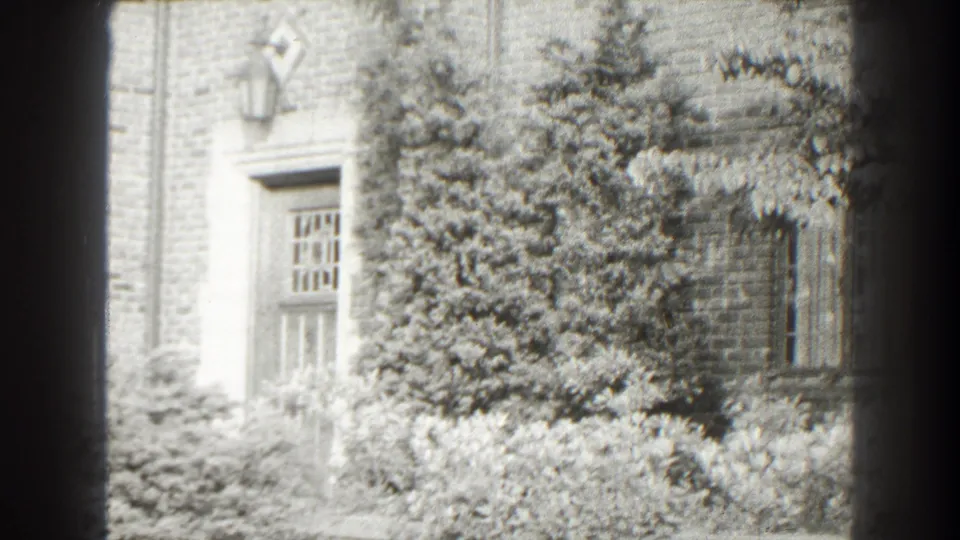 1938 home-movie view of a brick house facade with heavy ivy, a side doorway, barred-looking window panes, exterior lantern, and low shrubs.