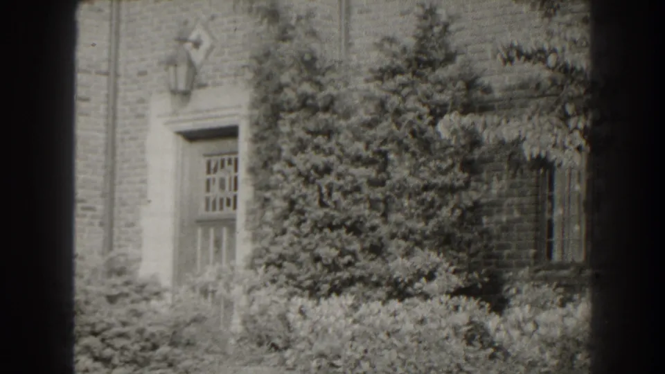 1938 Cumberland, Maryland 8mm frame showing a brick building entrance partly obscured by shrubs and a leafy tree.