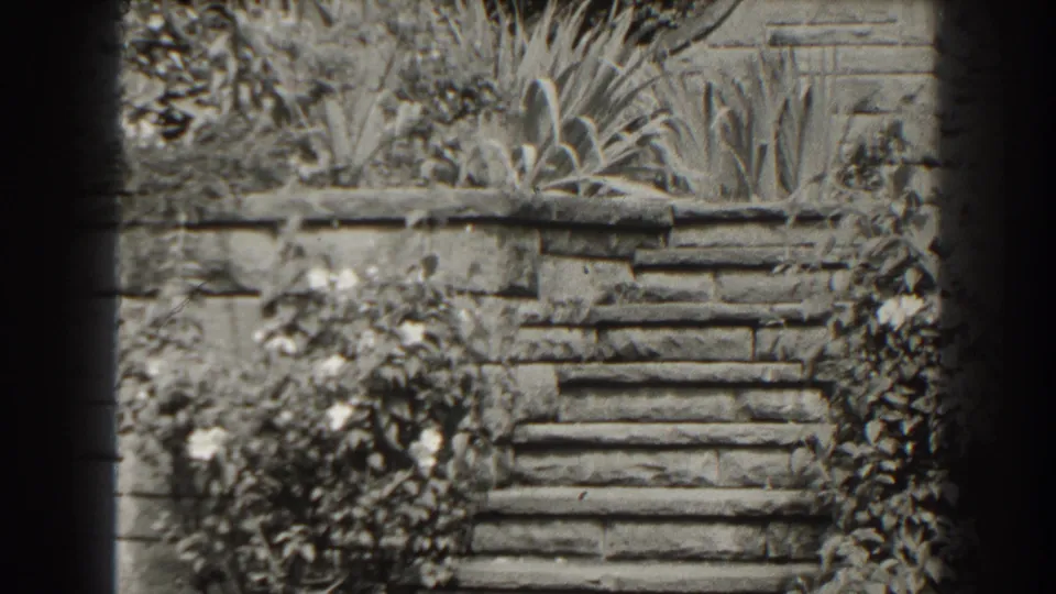 Black-and-white 1938 Cumberland frame with a low stone wall, steps, shrubs, and tall foliage filling the exterior view.