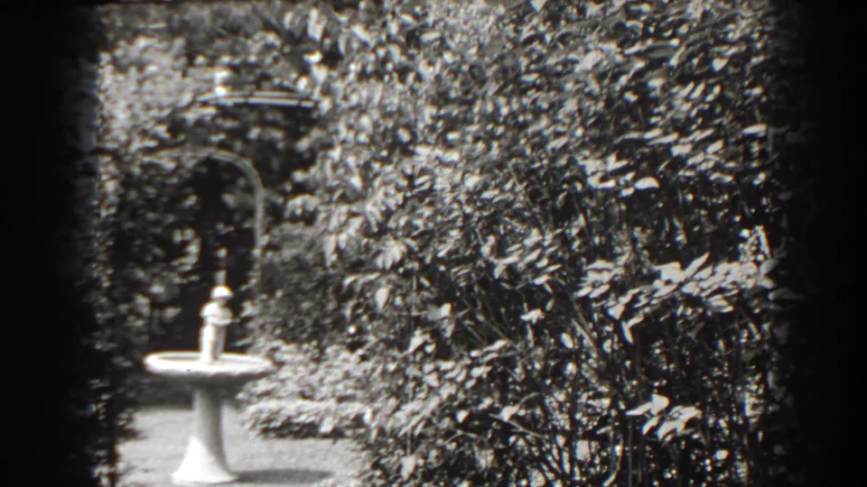1938 Cumberland, Maryland 8mm frame showing a white pedestal cherub fountain beside a path, with shrubs and an arbor-like feature behind it.