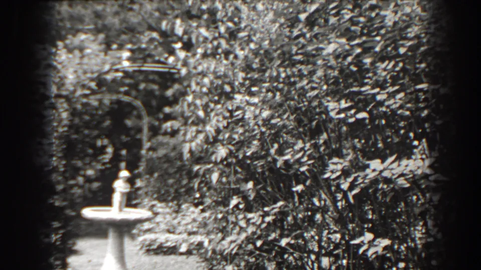 The closing frame holds on the white pedestal birdbath against dense shrubs and a shaded garden arch in Cumberland, Maryland.