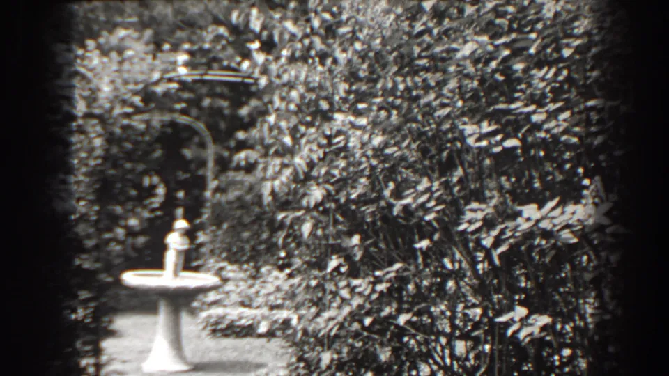 Black-and-white 1938 Cumberland garden view with a white bird bath, dense foliage, and soft 8mm frame edges.