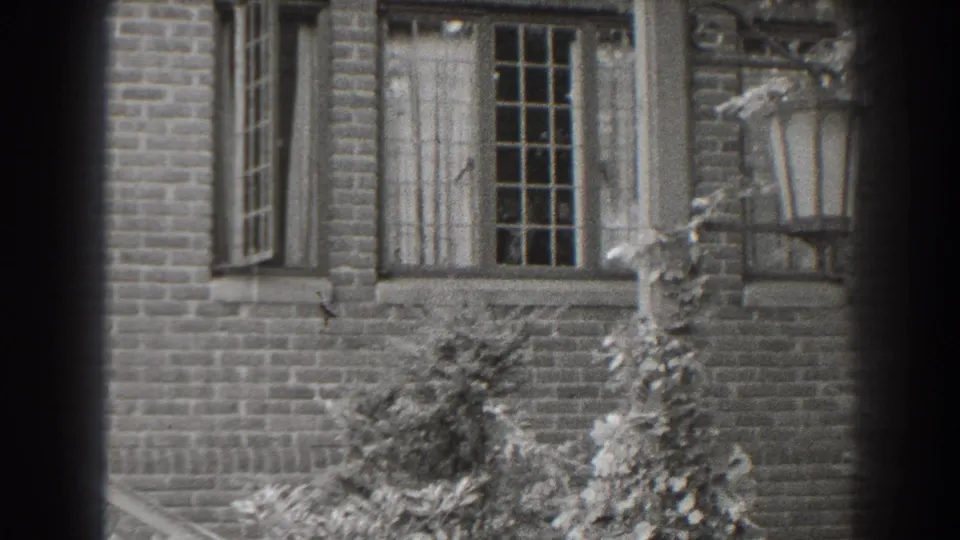 Black-and-white 1938 Cumberland home-movie frame of a brick exterior with a multi-pane window, shrubs, and lantern fixture.