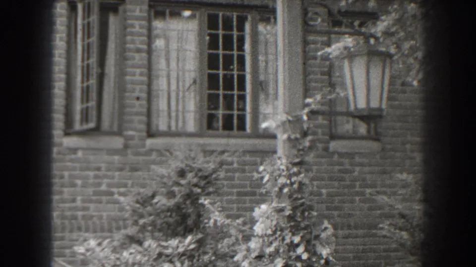1938 Cumberland, Maryland home-movie frame showing a brick house exterior with curtained windows, climbing vines and a mounted lantern.