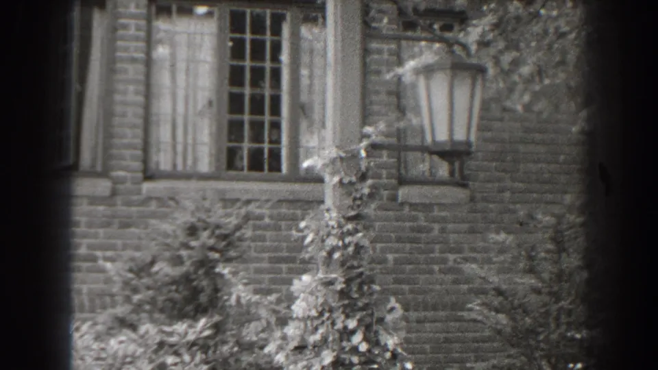 Black-and-white 8mm frame of a Cumberland brick house exterior with a tall gridded window, leafy vines and a lantern fixture.