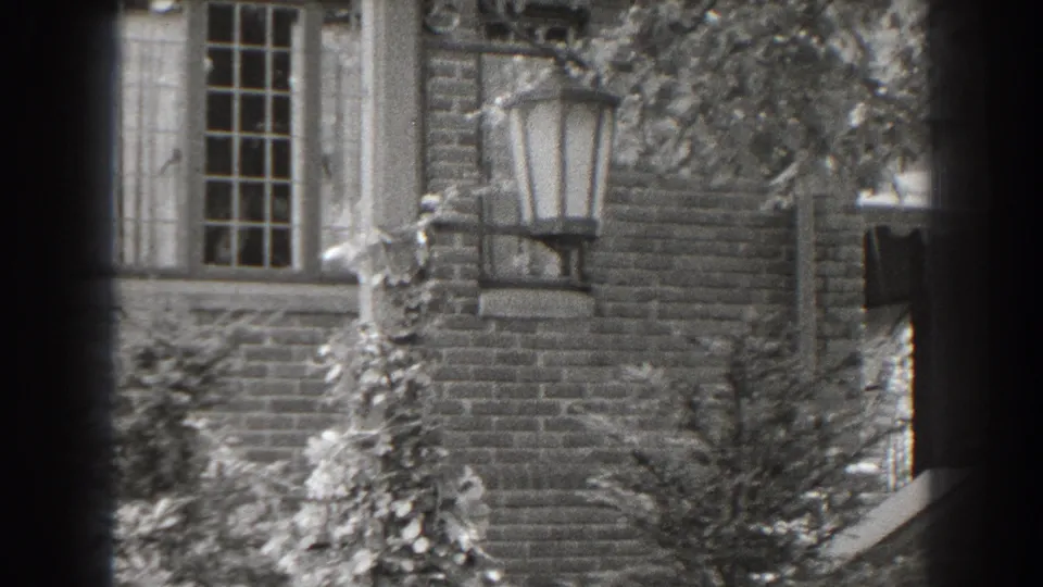 1938 home-movie still from Cumberland showing the brick facade, multi-pane window, lantern, climbing plants and garden foliage.