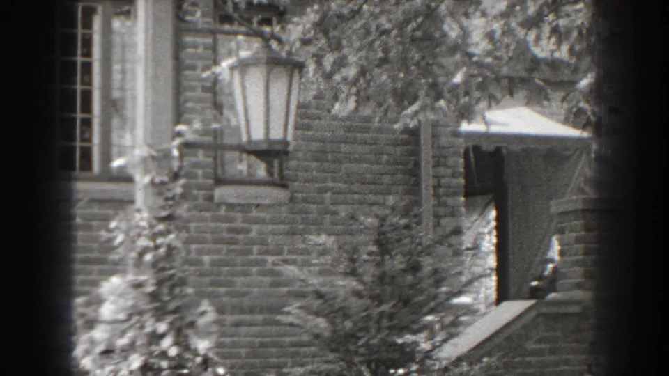 Black-and-white 8mm still of the Cumberland house exterior with brickwork, a window, vines, lantern and a partial awning or roof edge.