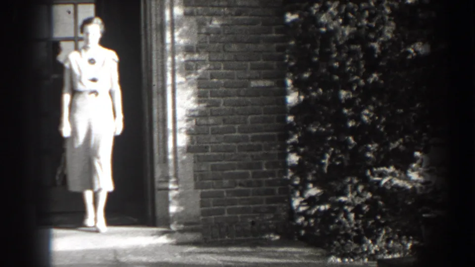In the 1938 Cumberland home movie, a woman in a light belted dress steps out from a brick house doorway onto the porch.