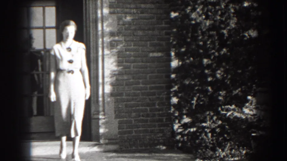 The woman continues walking toward the camera outside the Cumberland brick house, with shrubs and a window visible behind her.