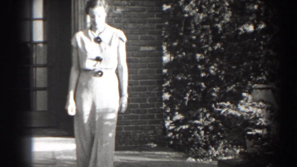 Adult woman walks toward the camera from a brick entrance, wearing a light dress with a dark belt and necklace.
