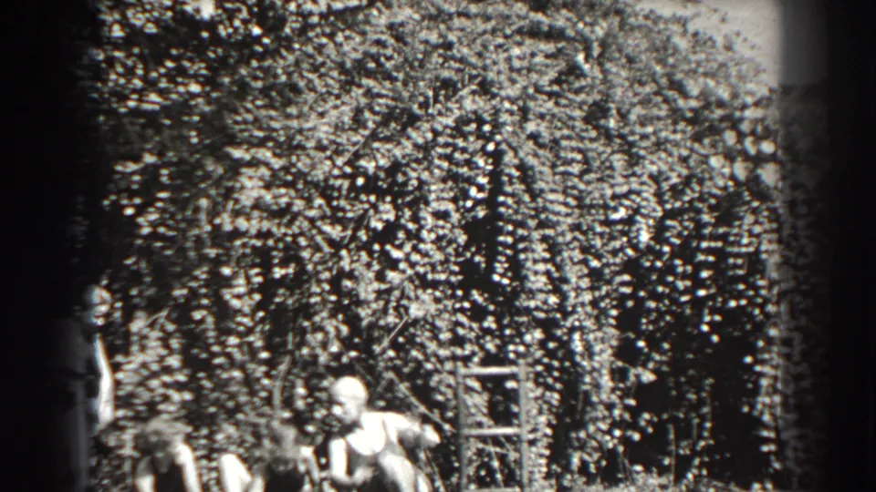 Black-and-white 1938 Cumberland frame with a seated woman in dark swimwear near dense bushes and a partial adult nearby.