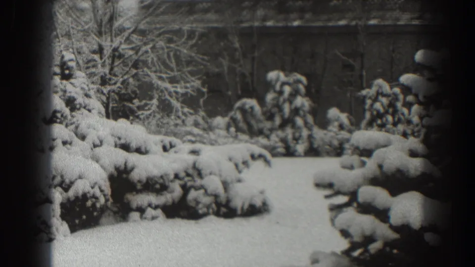 1938 Martinsburg home-movie frame showing snow-covered garden shrubs, bare branches, and a brick building beyond the lawn.