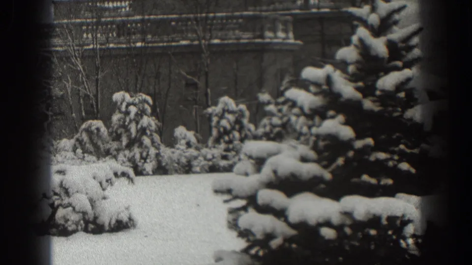 Late frame of the 1938 Martinsburg winter scene, with a snow-covered lawn, evergreen branches, bare trees, and brickwork behind.