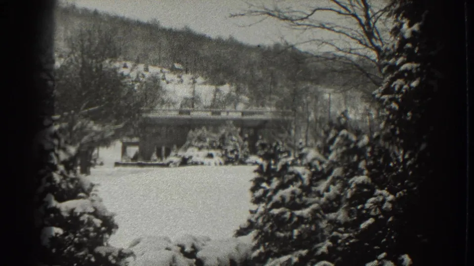 Black-and-white 8mm frame of a Martinsburg winter landscape, showing snow, a low house, hillside trees, and a bridge or raised roadway.