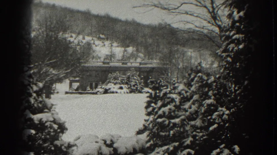 Martinsburg winter home-movie frame showing a snow-filled foreground, small house, hillside, and branches around the view.