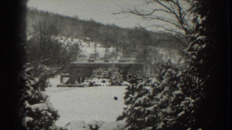 Late-1930s Martinsburg winter frame with snow-covered trees, a small house, hillside, and a dark speck in the film image.