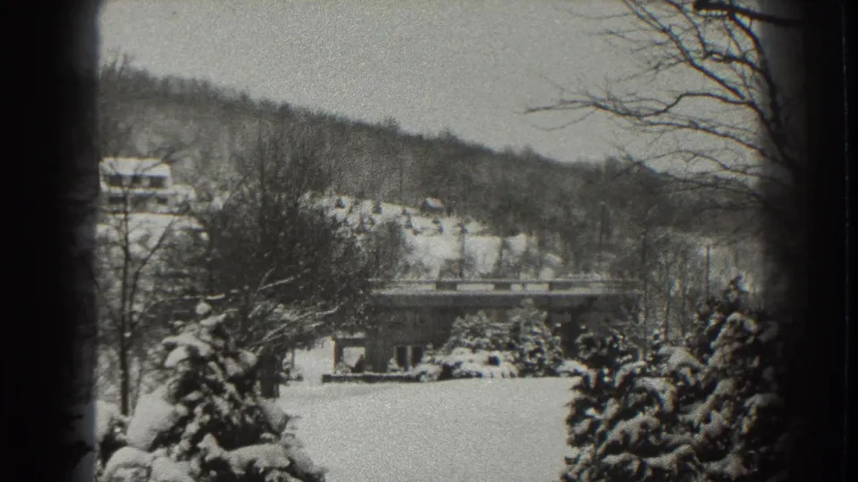 Home-movie frame of a snow-covered Martinsburg scene with foreground evergreens, a low bridge, and distant hillside buildings.