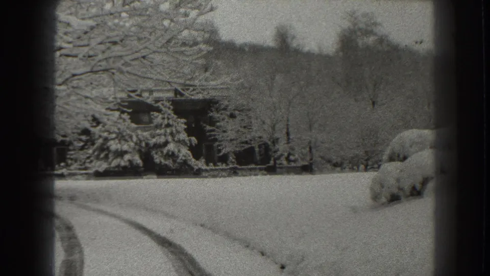 1938 Martinsburg, West Virginia home-movie frame showing snow across a rural yard, bare trees, shrubs, a small cabin, and road tracks.