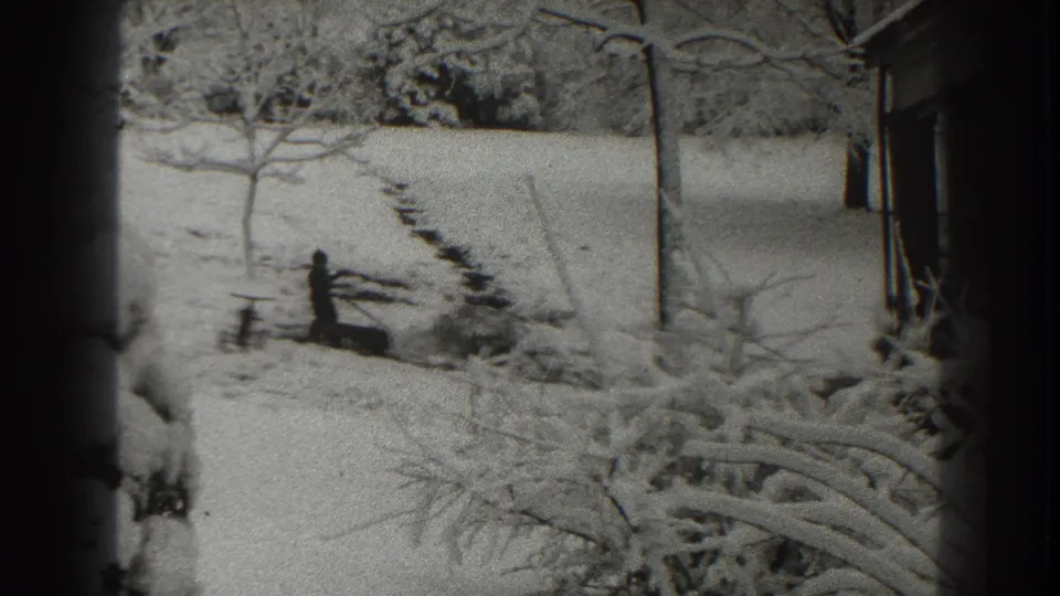 A man in dark clothing works with an axe near a log in the snowy Martinsburg yard, seen through grainy black-and-white 8mm footage.