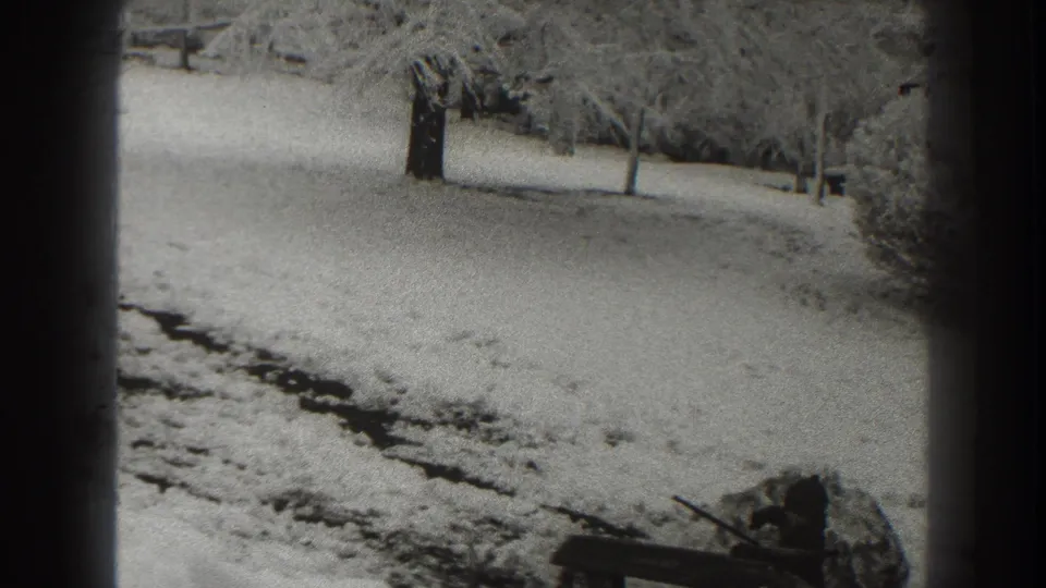 Black-and-white 8mm frame of a snow-covered Martinsburg landscape with trees, a dark track, and a wooden object near the lower right.