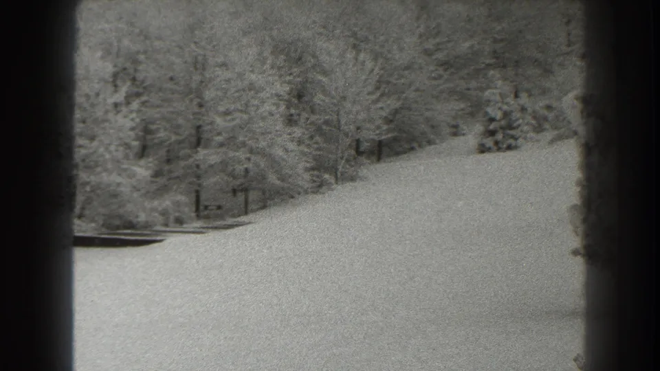 Black-and-white 8mm frame of a snow-covered Martinsburg slope, with bare trees, evergreens, and heavy dark framing at the side.