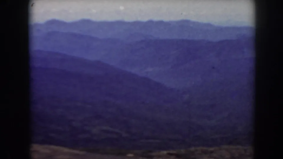 1938 home-movie frame of New Hampshire mountain slopes, softened by haze, grain, and a blue-purple color cast.