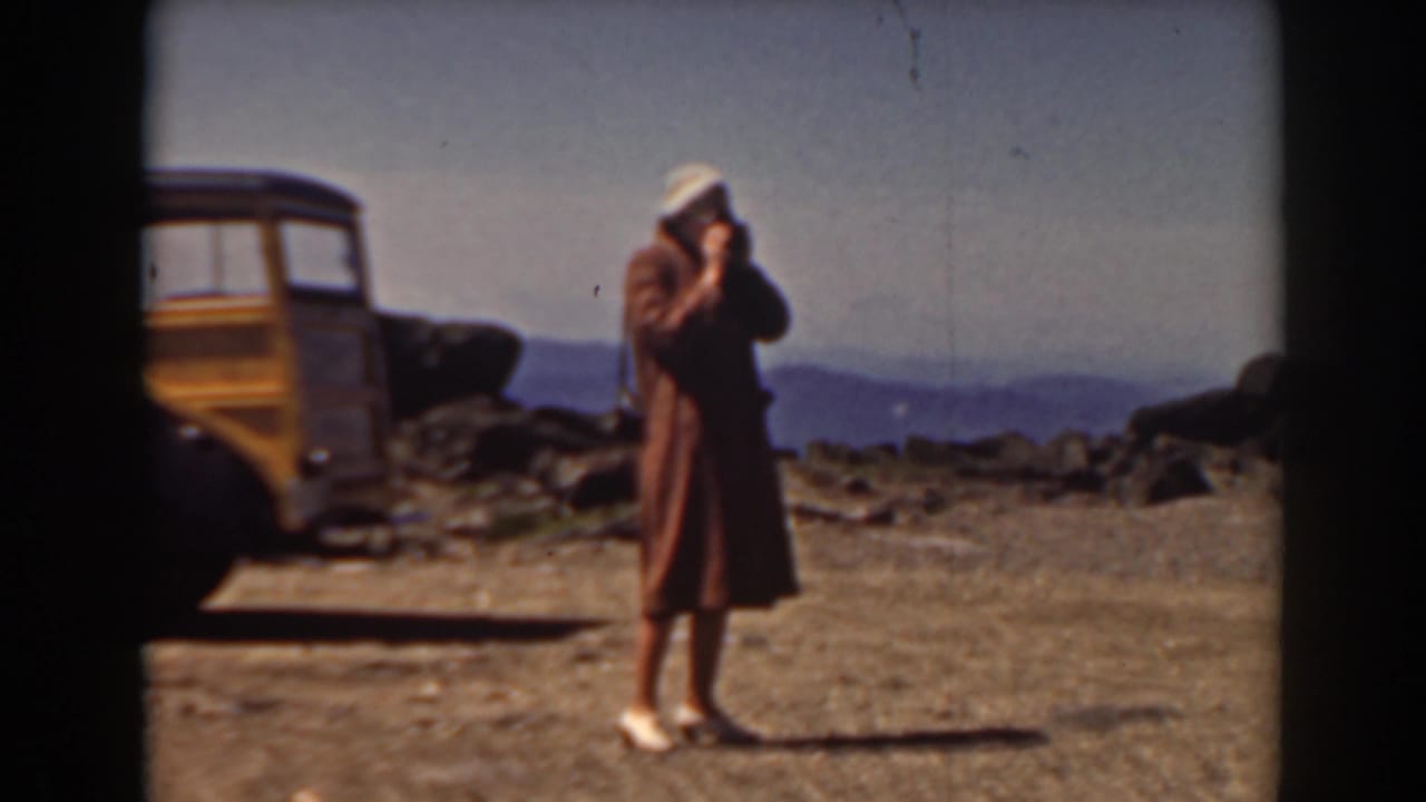 Woman in a brown coat photographs a rocky mountain summit near a wood-paneled station wagon.