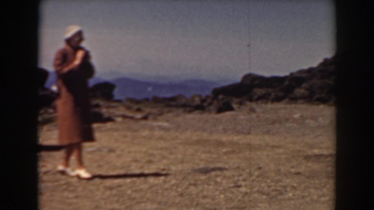 Woman with a camera stands on open rocky ground with distant mountain ridges behind her.