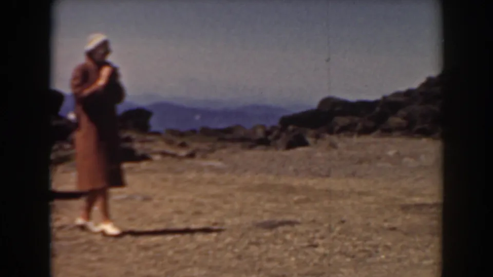 At the Mount Washington summit, the woman stands with a camera on gravelly terrain, with hazy mountain ridges in the background.