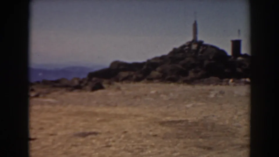 A wider Mount Washington summit view shows rocky ground, a raised marker on the peak, a small structure, and blue mountain ridges beyond.
