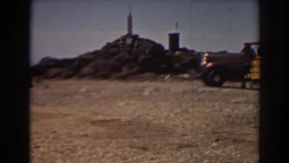 The Mount Washington summit marker and nearby structure remain in view, with part of a vintage vehicle entering the right edge of the frame.