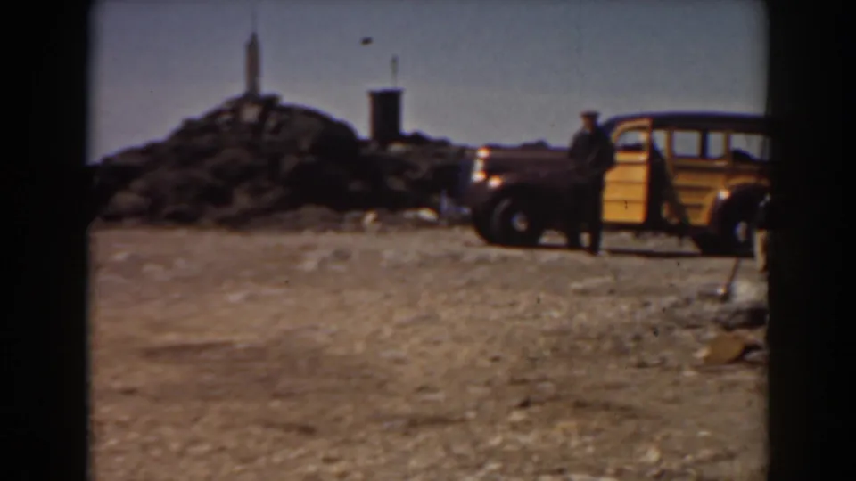 1938 Mount Washington home-movie frame showing a yellow wood-paneled car, a man near the door, rocky ground, and low summit terrain.
