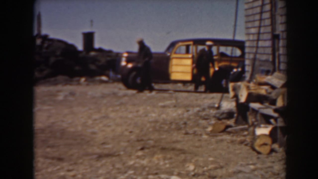 1938 Mount Washington frame with two adults around a yellow wood-paneled station wagon near a shingled building and rough ground.
