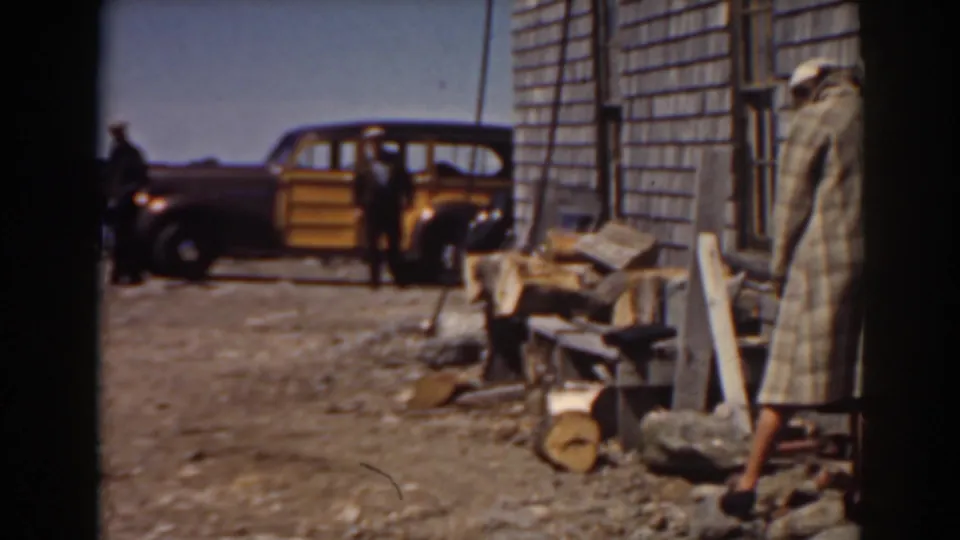 1938 Mount Washington home-movie frame showing the yellow car, a suited man, a long-coated adult, stacked firewood, and a shingled wall.