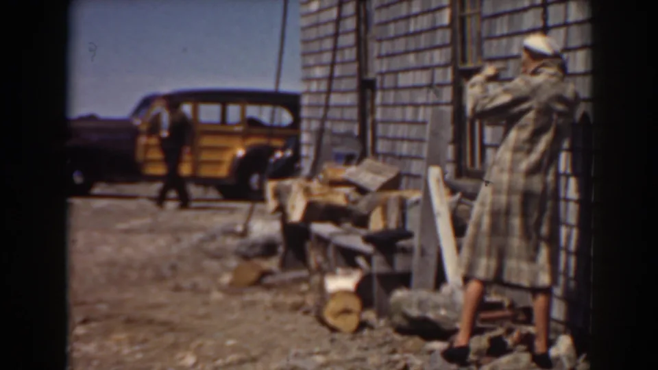 1938 Mount Washington frame with a long-coated adult near a shingled building, firewood in the foreground, and the yellow car behind.