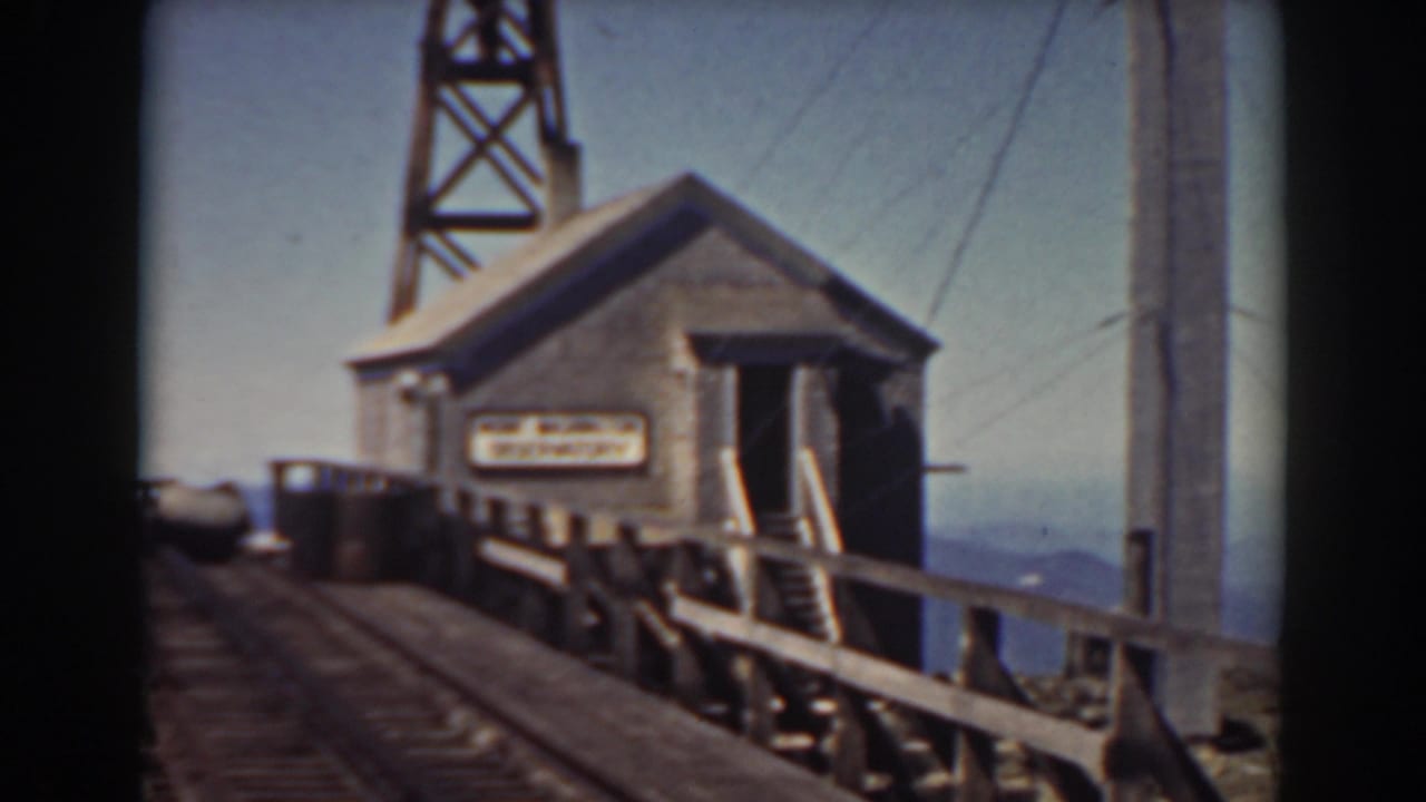 Small building beside railroad tracks on a bridge with a tower rising behind it.