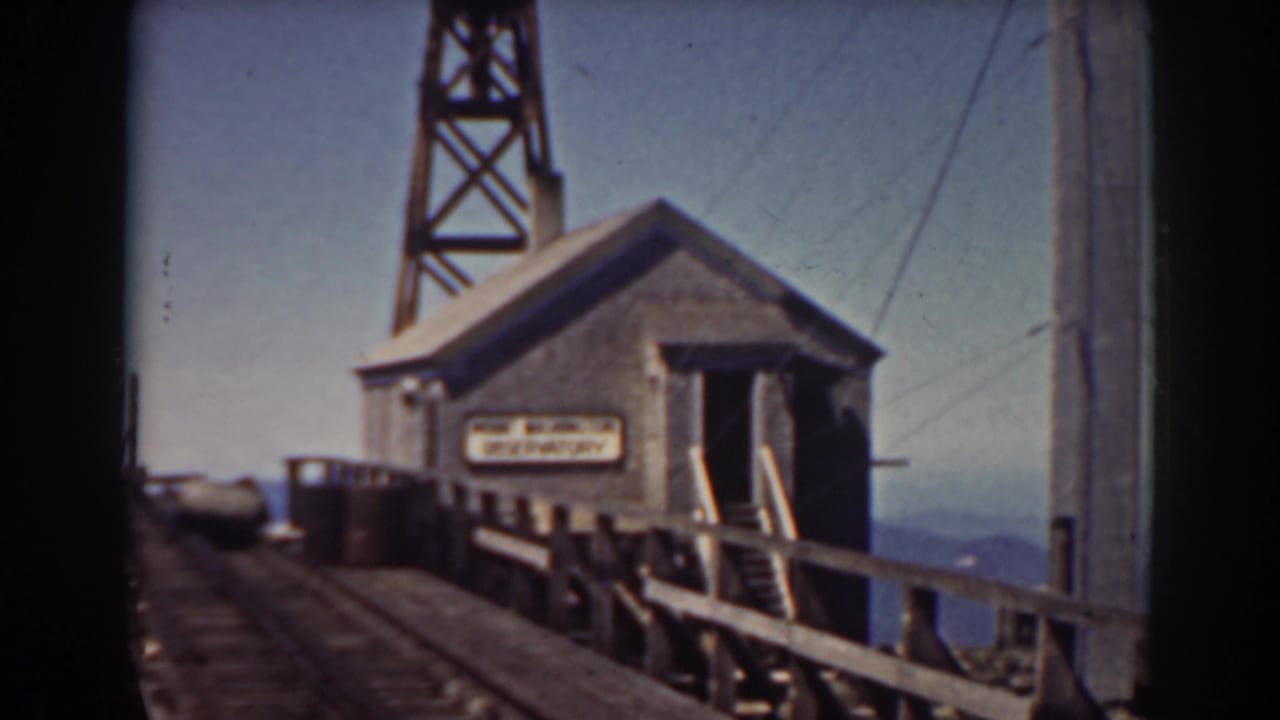 A small building on the Mount Washington railroad bridge appears beside rails and a railing, with a sign on the wall and tower framing the background.