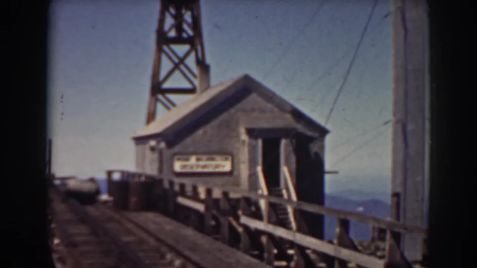 A small building on the Mount Washington railroad bridge appears beside rails and a railing, with a sign on the wall and tower framing the background.