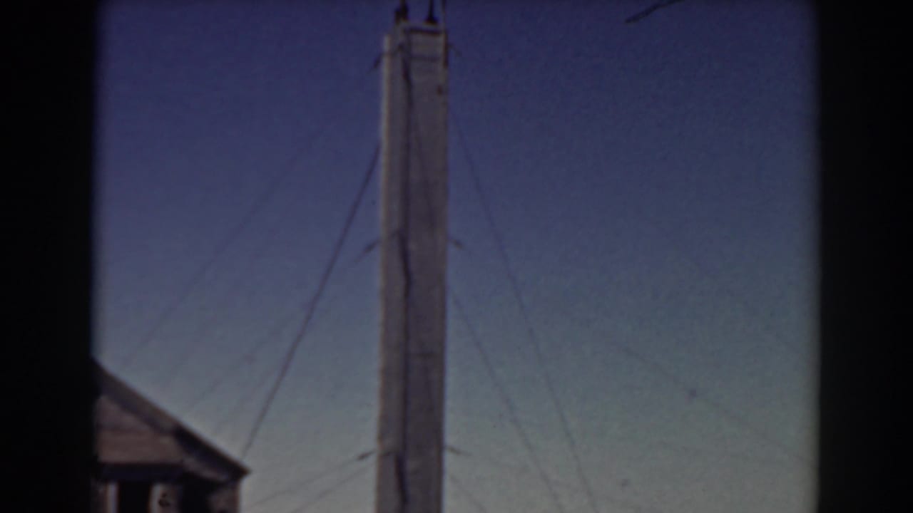 Tall wooden tower with guy wires against a blue sky, with a roof edge at the lower left.
