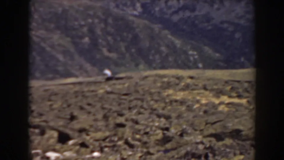 Mount Washington home-movie frame showing rough rocky ground, a distant hillside, and a small bright object in the landscape.