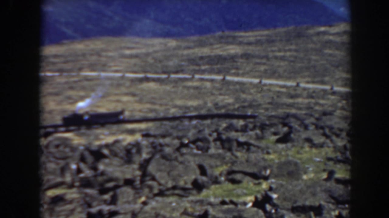 Steam locomotive backing along a curving mountain track with rocks and hillside behind it.